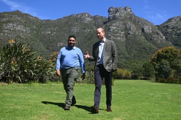 Two people walk on grass with mountains in the background.